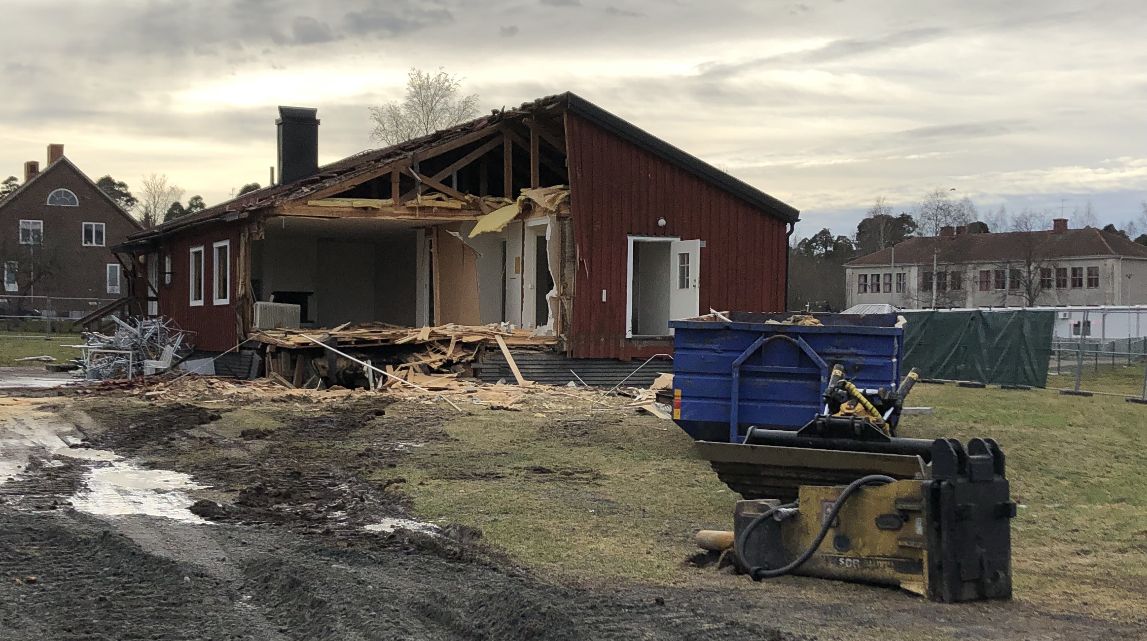 Hemgården rivs. Halva gavelväggen är borta. Container och maskindelar i förgrunden.