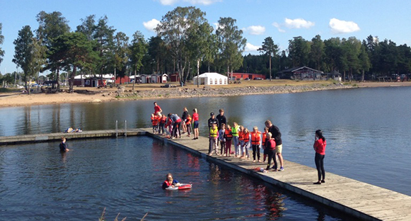 Hammarölägret 2019. Barn som står på bryggan vid Mörudden. Flytvästar på. Soligt väder. Ett barn simmar med räddningsringen.