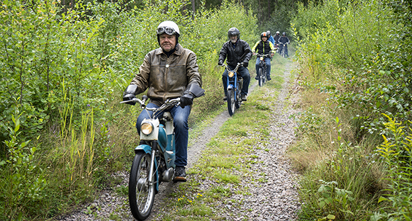 Äldre herrar som åker moped på skogsväg.